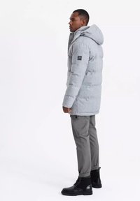 Parka gris, acolchada y con capucha, que presenta un tejido texturizado y un parche con el logo en la manga. Llevado con pantalones cargo grises y botas negras con cordones.