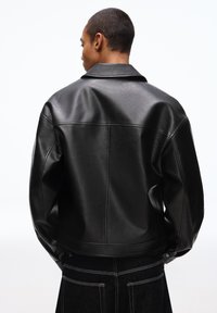 Person steht mit dem Rücken zur Kamera, trägt eine schwarze Lederjacke mit Kragen und eine schwarze Hose mit weißen Nähten auf den Gesäßtaschen.