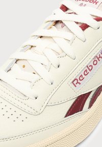 Κοντινό ενός κρεμ sneaker Reebok με λευκά κορδόνια, κόκκινο λογότυπο στο πλάι, διάτρητο μπροστινό μέρος και μπεζ υφή στην σόλα.