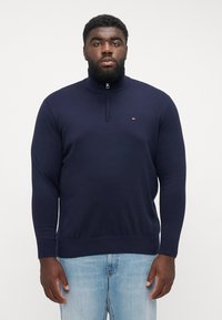 Granatowy sweter z zamkiem na szyi, wykonany z gładkiego materiału, z długimi rękawami, ściągaczami na mankietach i małym logo na klatce piersiowej. W połączeniu z jasnoniebieskimi dżinsami.