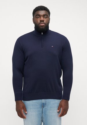 Pull à col zippé navy en tissu lisse avec des manches longues, des poignets côtelés et un petit logo sur la poitrine. Associé à un jean bleu clair.