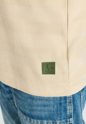 Roomkleurig shirt met een klein groen vierkant logo-patch bovenop een blauwe spijkerbroek op het onderlichaam van een persoon.