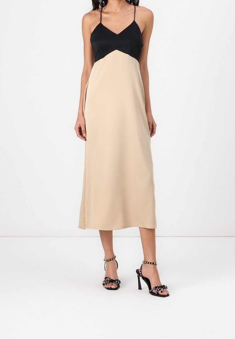 Femme portant une robe midi sans manches noire et beige avec des bretelles spaghetti et des sandales à talons hauts noires ornées de clous.
