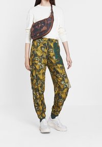Bloemmotieven, gele en groene cargobroek met geribbelde manchetten en zakken, gecombineerd met een witte trui en een bruine schoudertas.
