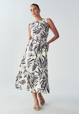 Vestido maxi en crema con estampado de hojas negras, con un corsé ajustado, cinturilla elástica y falda de volantes. Incluye tirantes finos y sandalias planas.