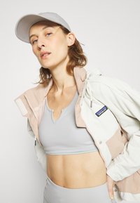 Casaco leve em blocos de cor, em bege e branco, combinado com uma blusa desportiva cinza. A textura é suave e o design apresenta um patch de logotipo visível. Um boné cinza completa o visual.
