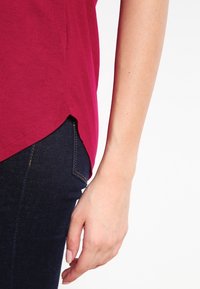 En kvindes hånd hviler ved siden af mørkeblå jeans og en rød skjorte med buet kant mod en hvid baggrund.