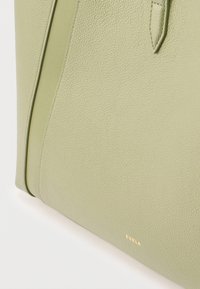 Κοντινό πλάνο γωνίας τσάντας από ανάγλυφο δέρμα ανοιχτού πράσινου χρώματος με χρυσή επιγραφή "Furla" και ραμμένη λεπτομέρεια ιμάντα.