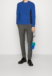 Maglione blu con pannelli laterali neri, pantaloni grigi su misura, scarpe nere stringate e una piccola borsa blu e verde.