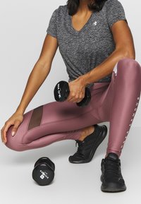 Grå träningsskjorta med v-ringad hals, rosa leggings med meshdetalj, svarta sneakers. En individ håller i en svart hantel märkt 3,0 kg.