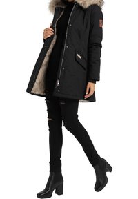 Schwarze Parka mit einer Kapuze aus Kunstfell, Reißverschluss vorne und Innenfutter. Mit einer Brusttasche und seitlichen Taschen ausgestattet. Kombiniert mit zerrissenen schwarzen Jeans und hohen Stiefeln.