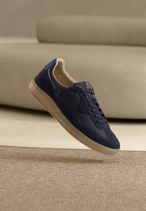 Scarpa da ginnastica blu navy con suola e rivestimento beige, sospesa sopra un tappeto beige testurizzato, con divano beige sullo sfondo.