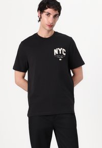 T-shirt en coton noir à manches courtes. Présente des graphismes et du texte blancs "NYC" sur la poitrine. Encolure classique, coupe décontractée.