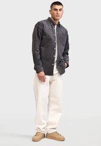 Dunkelgraues Kordhemd mit Button-down-Kragen, Brusttasche, kombiniert mit cremefarbigen Baumwollhosen und beigen Sneakers. Einfarbiger Hintergrund.