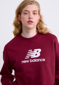Sweatshirt bordeaux avec un col rond, arborant un logo "new balance" blanc et des rayures, fabriqué à partir d'un tissu doux avec une coupe décontractée.