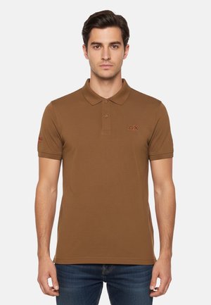 Uomo che indossa una polo marrone a maniche corte con colletto e patta con bottoni, abbinata a jeans blu, in piedi contro uno sfondo semplice.