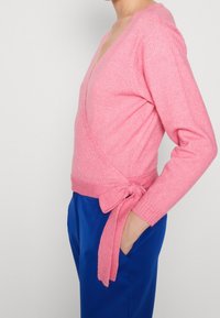 Růžový svetr s křížením vyrobený z měkké pleteniny. Má hluboký výstřih ve tvaru V, dlouhé rukávy a detail uvázání v pase. Nosí se s modrými kalhotami.
