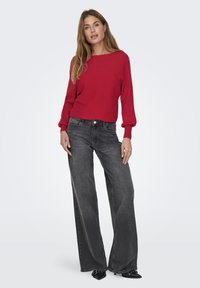 Red ribbed sweater with wide boat neckline and puffed sleeves paired with high-waisted, dark gray wide-leg jeans and pointed black shoes.
