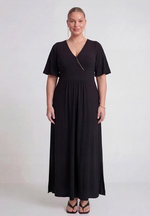 Frau steht und trägt ein schwarzes knöchellanges Kleid mit flatternden Ärmeln, V-Ausschnitt, goldenen Schmuck und schwarzen Sandalen vor einfarbigem Hintergrund.