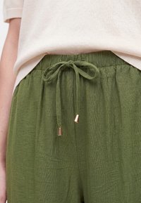 Green loose-fitting pants with a drawstring waist tied in a bow, worn with a light beige top partially visible above the waistband.