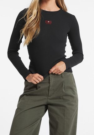 Donna che indossa una maglia aderente a maniche lunghe nera con il logo rosso "Guess Jeans" e pantaloni verde oliva a vita alta, con le mani che sistemano la maglia.
