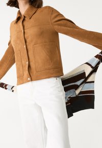 Veste en daim marron avec un col, des boutons-pression et deux poches avant ; pantalon blanc et un vêtement en tricot rayé drapé sur le bras.