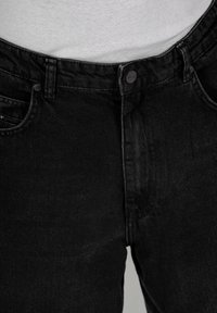 Los jeans de mezclilla negra presentan un diseño clásico con cierre de cremallera, dos bolsillos frontales y una textura sutil. La tela parece ligeramente desgastada.