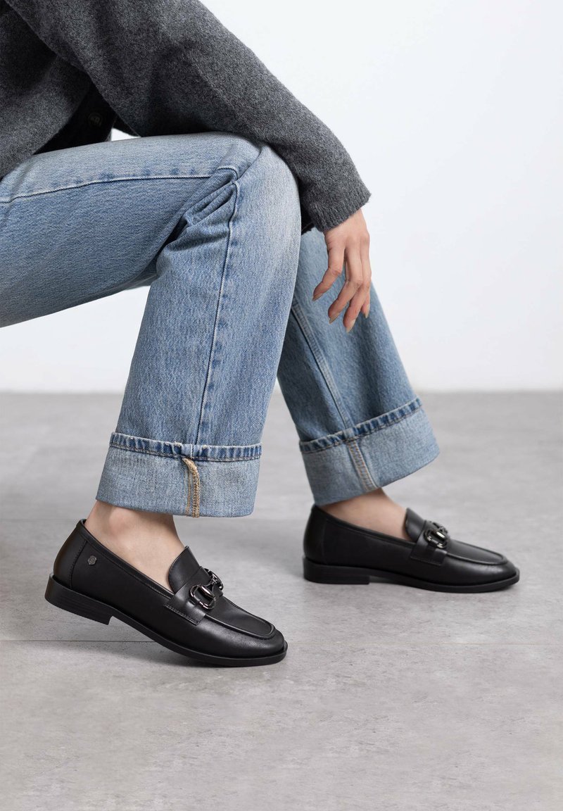 Mocasines de cuero negro con un hebilla decorativa, con punta redondeada y tacón bajo, combinados con jeans de dobladillo enrollado en azul claro.