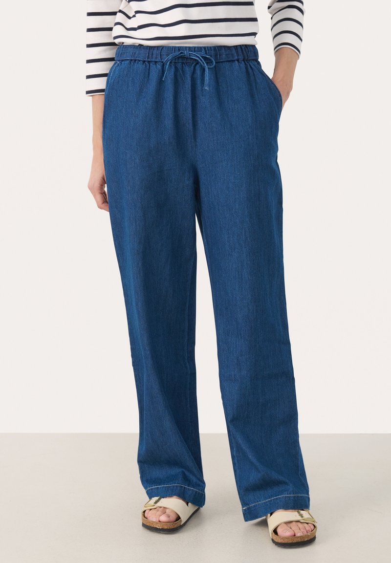 Femme portant un pantalon en denim bleu ample avec une taille élastique et un cordon de serrage, associé à des sandales blanches et un haut à manches longues rayé.