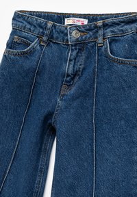 Siniset farkut, joissa on leveät lahkeet, viisi taskua, näkyvät saumat ja metallinen nappisulku vyötäröllä.