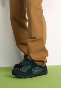 Čierne a tyrkysové turistické topánky s textúrovanou látkou a gumovou podrážkou, so šnúrkovým dizajnom, nosené s béžovými nohavicami na zelenej podlahe.