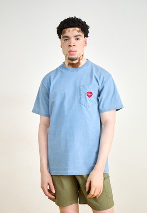 T-shirt en coton bleu clair à manches courtes, présentant une poche poitrine ornée d'un logo de cœur rouge. Associé à un short vert olive.