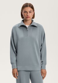 Svetlosivý pulóver s polozipsovým golierom, mäkká tkanina, voľný strih, rebrované akcenty na manžetách a leme, a textúrovaný panel na hrudi.