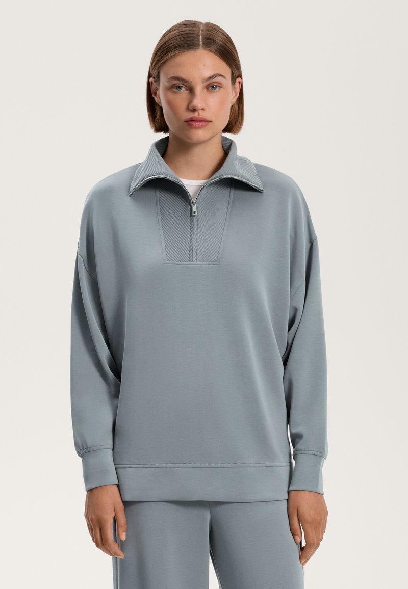 Svetlosivý pulóver s polozipsovým golierom, mäkká tkanina, voľný strih, rebrované akcenty na manžetách a leme, a textúrovaný panel na hrudi.