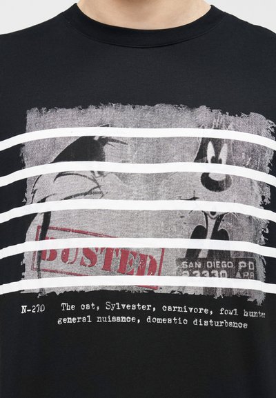 Chemise noire avec un design de type photo d'identité représentant le chat cartoon Sylvester derrière des barreaux blancs, tampon rouge "ARRÊTÉ", et texte décrivant les traits du personnage.