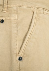 Beżowe spodnie bawełniane z przednią kieszenią ozdobioną srebrnym metalowym guzikiem, widocznymi stębnowaniami oraz miękkim, teksturowanym wykończeniem.