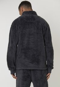 Homme portant une veste en polaire grise foncée et un pantalon assorti, vu de dos sur un fond clair et uni.