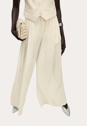 Persona dalla pelle scura che indossa pantaloni larghi color crema e un gilet, tiene in mano una pochette intrecciata color crema, indossa scarpe appuntite argento e un braccialetto d'argento.