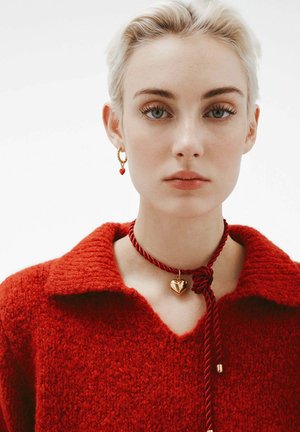 Mujer con cabello rubio corto que lleva un suéter texturizado rojo, un collar de cuerda roja con un colgante de corazón dorado y pendientes de aro dorados con encantos en forma de corazón rojo.
