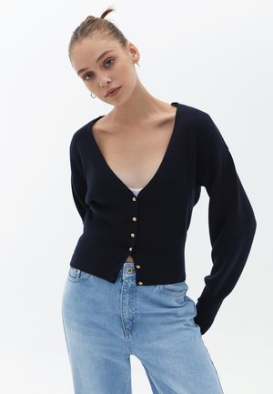 Marinefarbener V-Ausschnitt-Cardigan mit langen Ärmeln, goldenen Knöpfen an der Vorderseite und taillierter Passform. Kombiniert mit hellblauen Denim-Jeans.