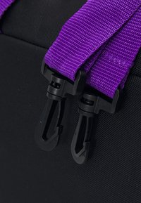 Fekete textil táska, jellegzetes lila nylon pántokkal, műanyag csatokkal. A pántok textúrázott szövésmintát mutatnak.