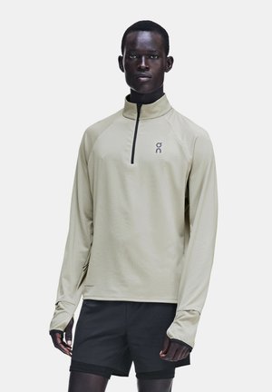 Homme portant un haut de sport beige clair à manches longues avec demi-fermeture éclair et un short noir, debout devant un fond clair uni.