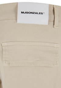 Beige szövet, texturált felülettel, hátsó zsebbel és egy fehér címkével, rajta a "MJGONZALES" felirattal fekete szöveggel.