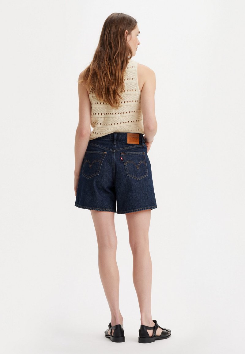 Shorts en denim bleu foncé, avec une coupe droite, arborant une petite étiquette en cuir et des coutures classiques sur les poches arrière. Portés avec un haut en tricot.