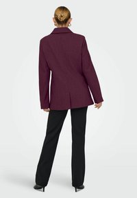 Bordeauxroter Blazer mit strukturierten Schultern, einfacher Rückenbelüftung und scharfen Revers, kombiniert mit schwarzen Hosen und hohen Schuhen, von hinten betrachtet.