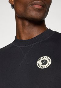 Sudadera de forro polar en color navy con cuello de canalé. Presenta un parche circular con un zorro negro y el texto "FJÄLLRÄVEN SWEDEN 1960". Textura suave.