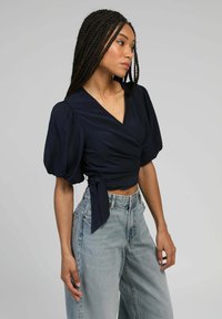 Femme avec des cheveux tressés portant un haut enroulé bleu marine avec des manches bouffantes et un jean taille haute bleu clair, se tenant devant un fond uni.