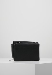 Kompaktní černá crossbody taška s texturovaným povrchem, stříbrným zipem a kovovými doplňky, odpočívající na bílém úhlovém podstavci na světle šedém pozadí.