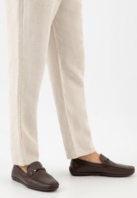 Braune Lederloafer mit glatter Oberfläche, kontrastierenden Nähten und silbernen Hardware-Details. Getragen mit hellbeigen Leinenhosen.
