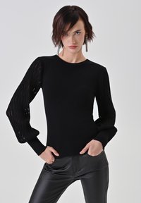 Schwarzes Oberteil mit langen, strukturierten, voluminösen Ärmeln, die vertikale Ausschnitte aufweisen. Enganliegende Form mit rundem Ausschnitt, kombiniert mit schwarzen Hosen.